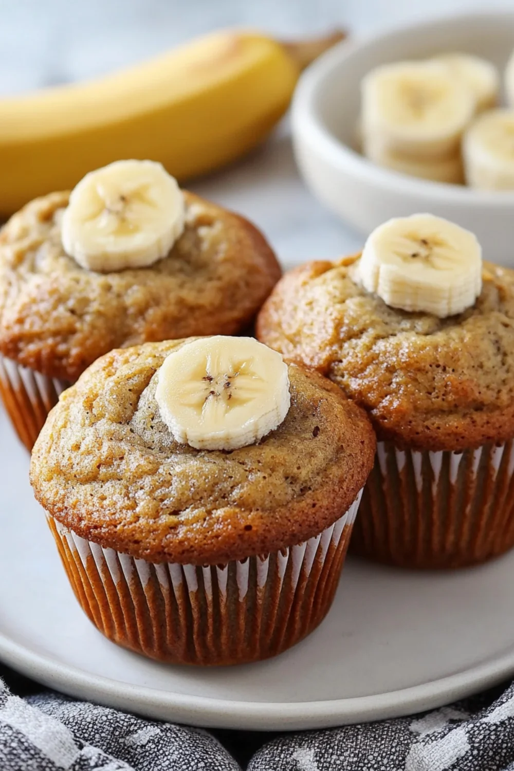 A close-up image of freshly baked banana muffins topped with banana slices, placed on a plate with a banana in the background.